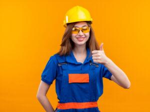 young-woman-builder-worker-construction-uniform-safety-helmet-smiling-broadly-showing-thumbs-up-standing-orange-wall
